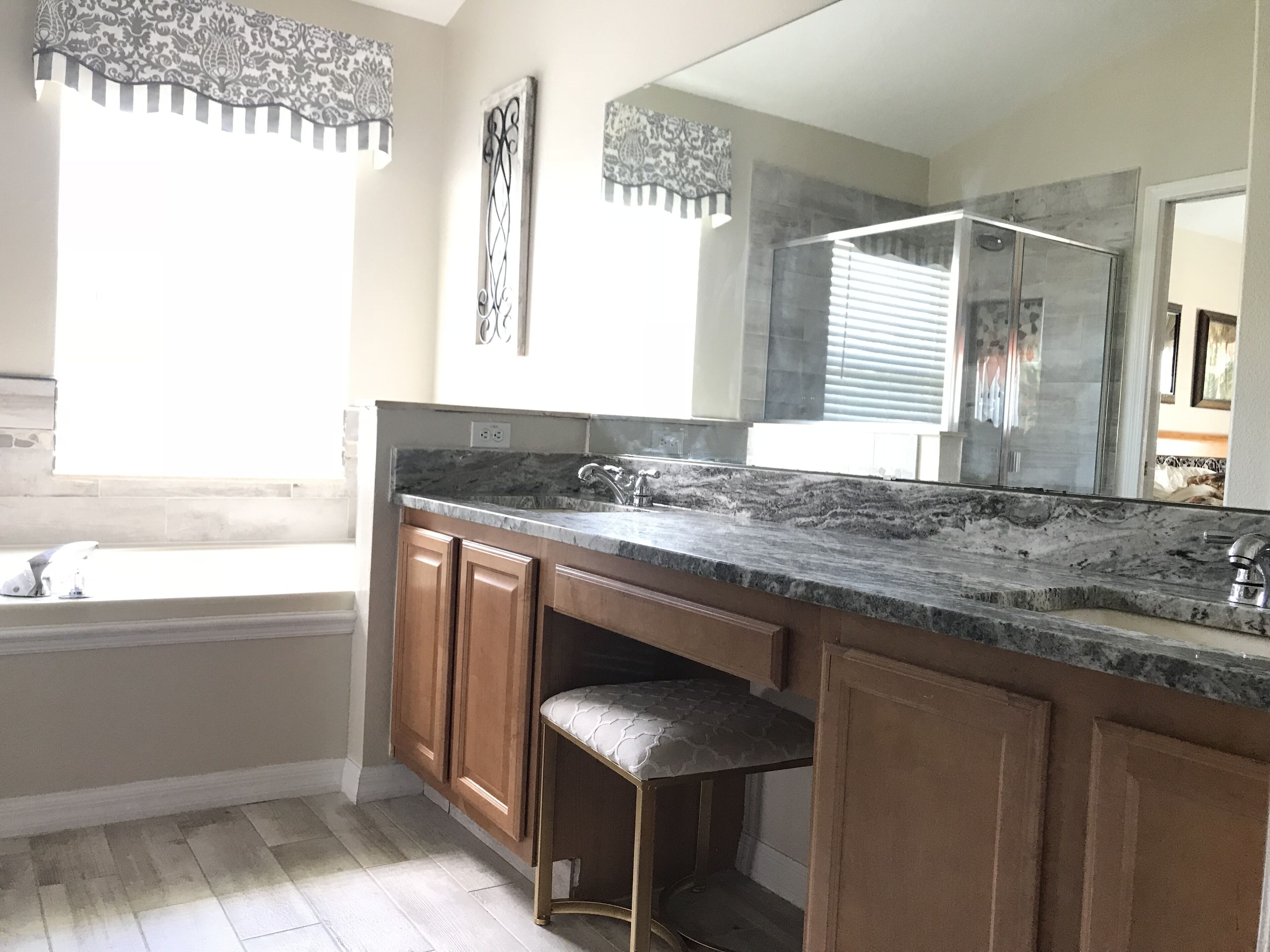 A modern bathroom with a large mirror, granite countertop, wooden cabinets, and a window with patterned curtains.