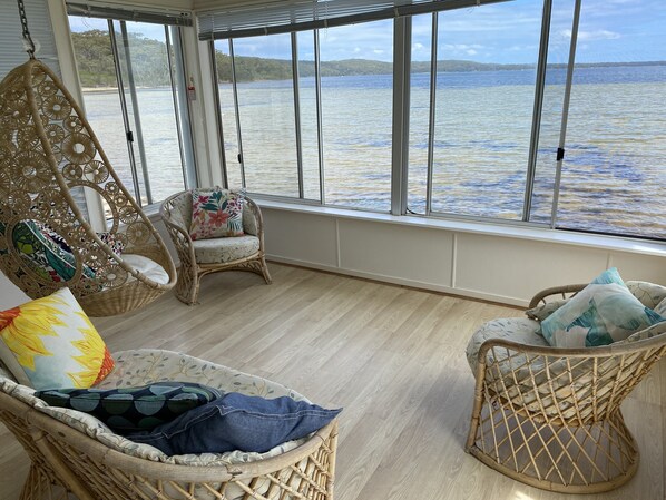 A room with wicker chairs, a hanging chair, and a view of a lake. - The Palm Tree Cottage - Absolute Waterfront - 5 mins to Hyams Beach (Erowal Bay)
