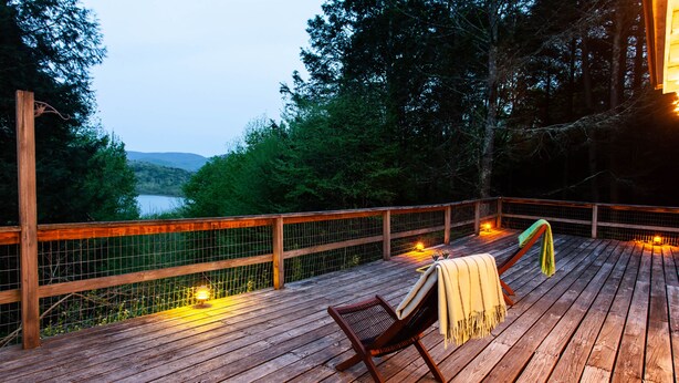 Une terrasse en bois avec une chaise et une couverture, d’où l’on a vue sur un lac et des collines boisées.