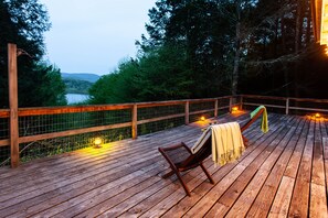 A wooden deck with a chair and a blanket, overlooking a lake and forested hills.