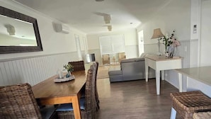 Uma sala ampla com uma mesa de jantar de madeira, cadeiras de vime, um sofá cinza e uma mesa de centro branca.