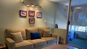 A living room with a beige sofa, a wooden coffee table, and a glass door leading to a balcony. - Modern, remodeled, ocean views, comfortable and convenient 580 sf (Kihei)