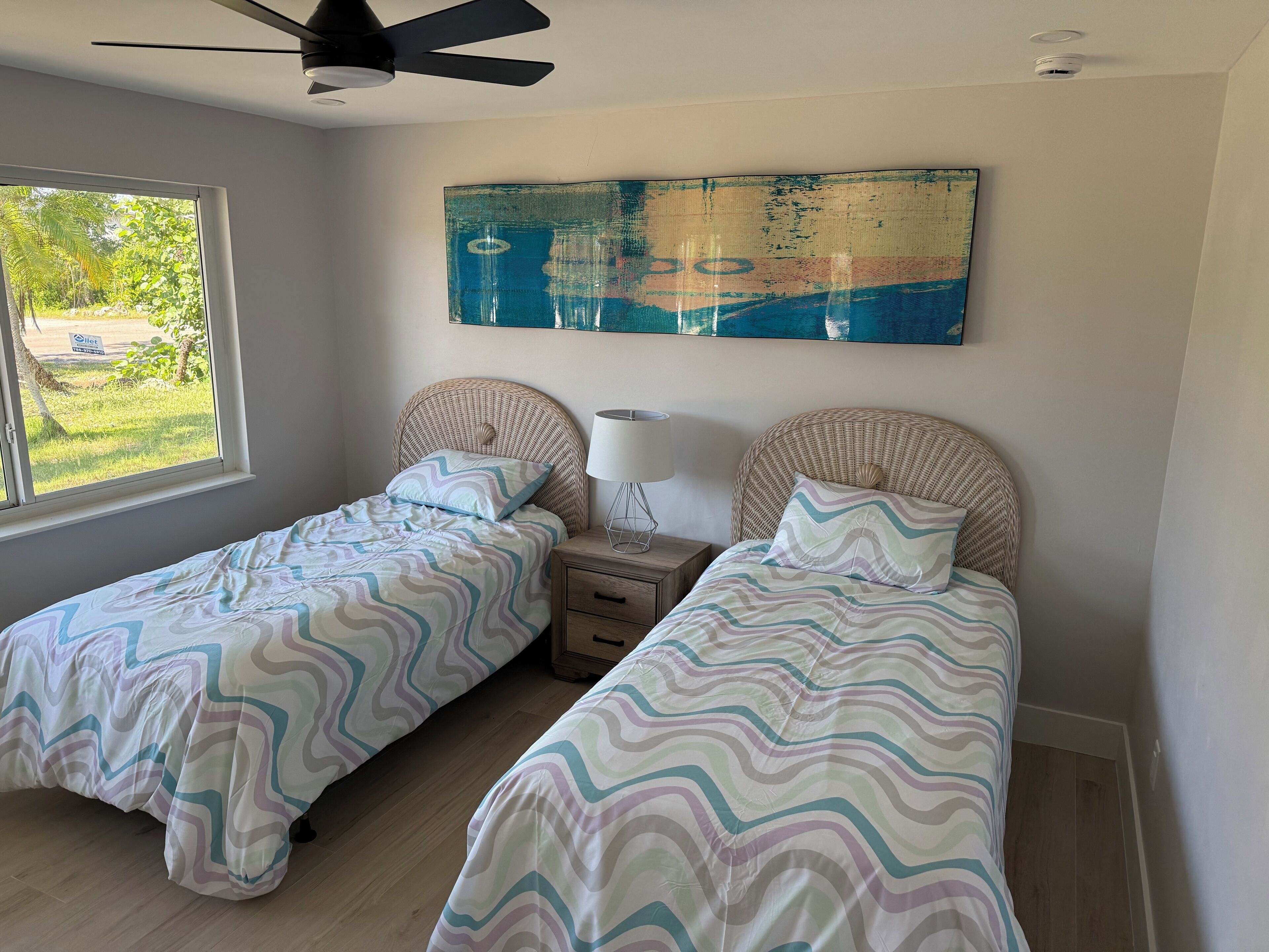 Une chambre avec deux lits simples, un ventilateur de plafond, un grand tableau abstrait et une fenêtre donnant sur un paysage verdoyant.