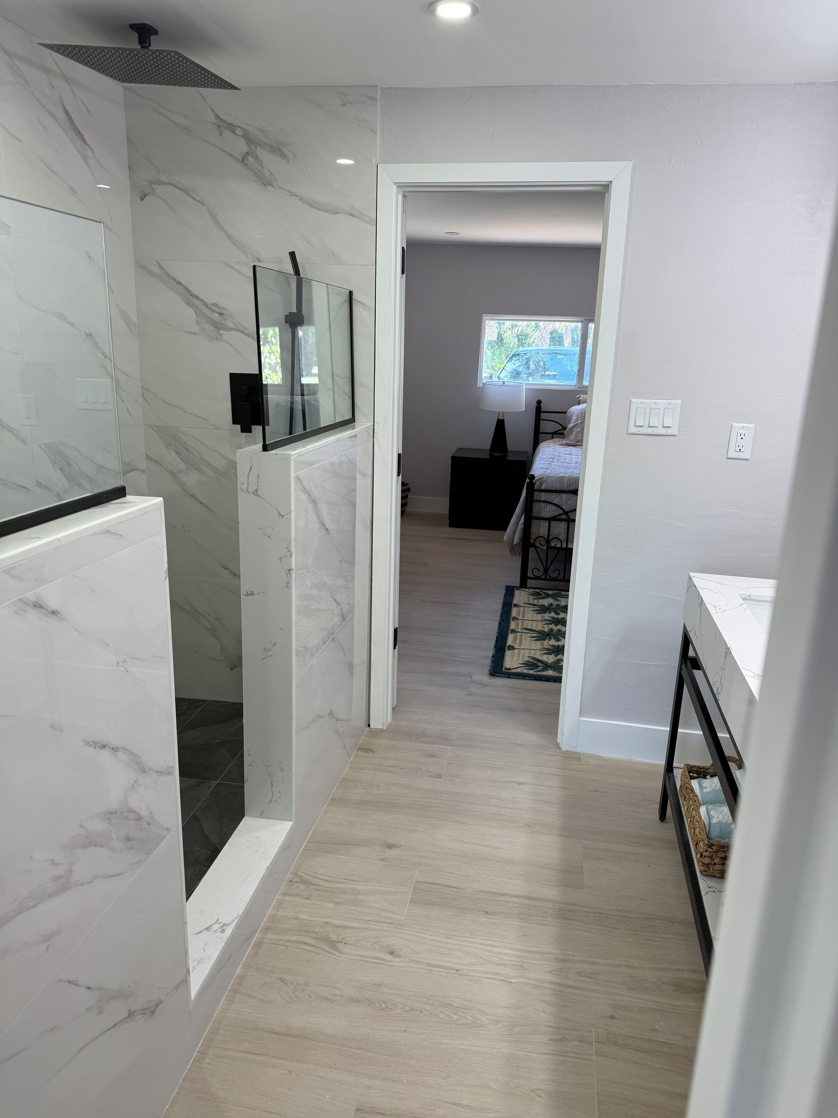 Une salle de bain moderne avec une douche, un miroir et des toilettes. Un couloir mène à une chambre à coucher équipée d’un lit et d’une fenêtre.