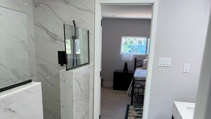 Uma casa de banho moderna com chuveiro, espelho e sanita. Um corredor leva a um quarto com cama e janela.