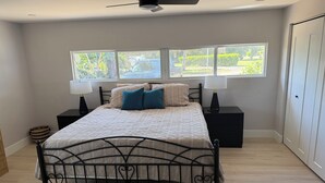 Quarto com cama, duas mesas de cabeceira, ventilador de teto e janela com vista para área verde.