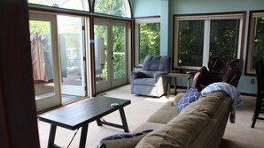 A living room with a sofa, two armchairs, a coffee table, and a small table with a lamp. There are large windows providing a view of the outdoors. - BEACHFRONT /HOTTUB/STEAM ROOM/GAME ROOM/JACUZZI/PETS/MASSAGE CHAIR/KAYAKS/BIKES (Michigan City)