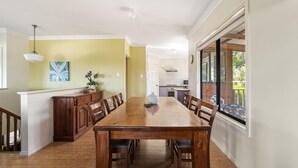 A dining area with a wooden table and chairs, a kitchen in the background, and a view of the outdoors through large windows. - Waterside View - A family-friendly retreat - Waterside View - family-friendly retreat with stunning Blackwood River views (Augusta)