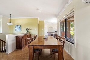 A dining area with a wooden table and chairs, a kitchen in the background, and a view of the outdoors through large windows.