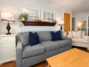 Un salon avec un canapé gris, deux coussins bleus, une table basse en bois, une table d’appoint blanche avec une lampe, et un fauteuil blanc.