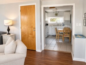 A room with a wooden door, a dining table with chairs, a kitchen area with cabinets, and a lamp on a bedside table.