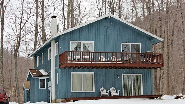 Et tosset etage blåt hus med balkon, skorsten og to Adirondack-stole udenfor.