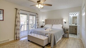 A bedroom with a large bed, a ceiling fan, a window with curtains, a bench, and a bathroom with a mirror and sink. - "Sunny Days"-Relaxation for All! Wifi-4 HDTVs-Open/Cov Dock-Game Rm-Linens Incl (Moneta)