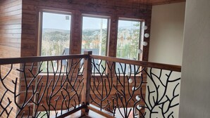 Een houten balkon met een metalen hekwerk, grote ramen en uitzicht op een bergachtig landschap.