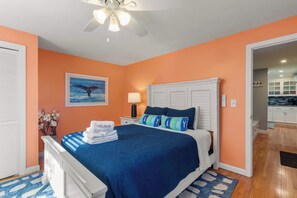 A bedroom with a bed, a ceiling fan, a nightstand, a lamp, a framed picture of a whale tail, and a bathroom visible through an open door.