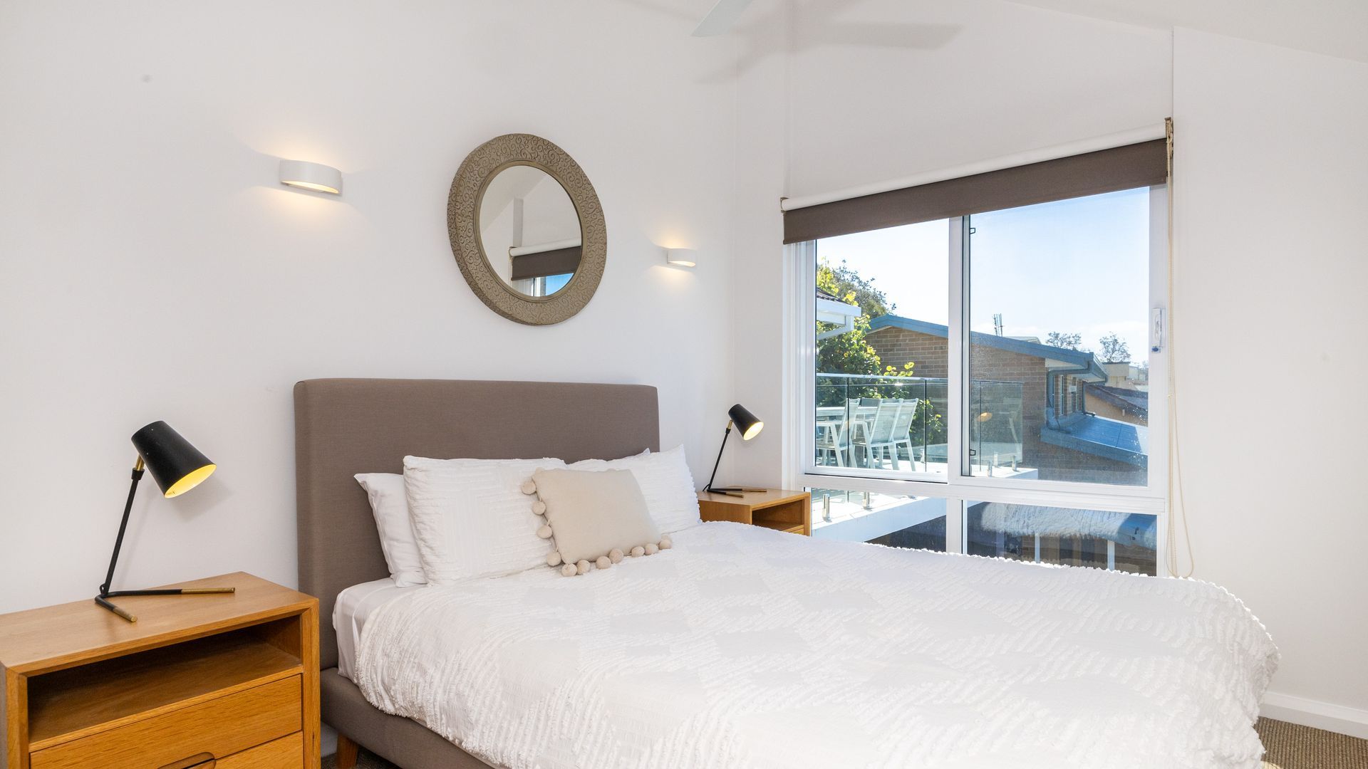 A bedroom with a large bed, two bedside tables, a round mirror, and a large window with a view of the outside.
