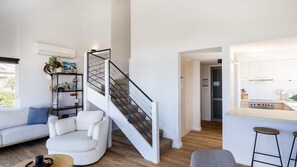 Un intérieur moderne comprenant un escalier, un espace de vie avec un canapé et un fauteuil, une cuisine équipée d’une cuisinière, et un coin bar avec des tabourets.