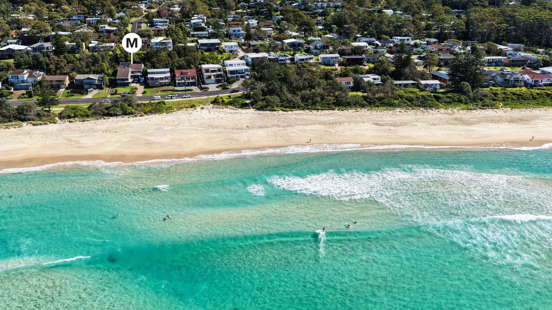 An aerial view of a coastal residential area with houses, a beach, and clear turquoise waters.