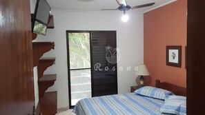 Quarto com cama, mesa de cabeceira, abajur, ventilador de teto, televisão fixada na parede e porta deslizante para uma varanda.