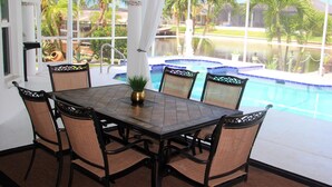 Un coin repas avec une table à plateau en verre, six chaises en osier et une vue sur une piscine et des palmiers.