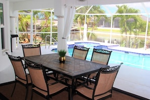 Un comedor con una mesa de vidrio, seis sillas de mimbre y vista a una piscina y palmeras.