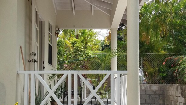 A covered porch with a white railing, surrounded by lush greenery and a stone wall.