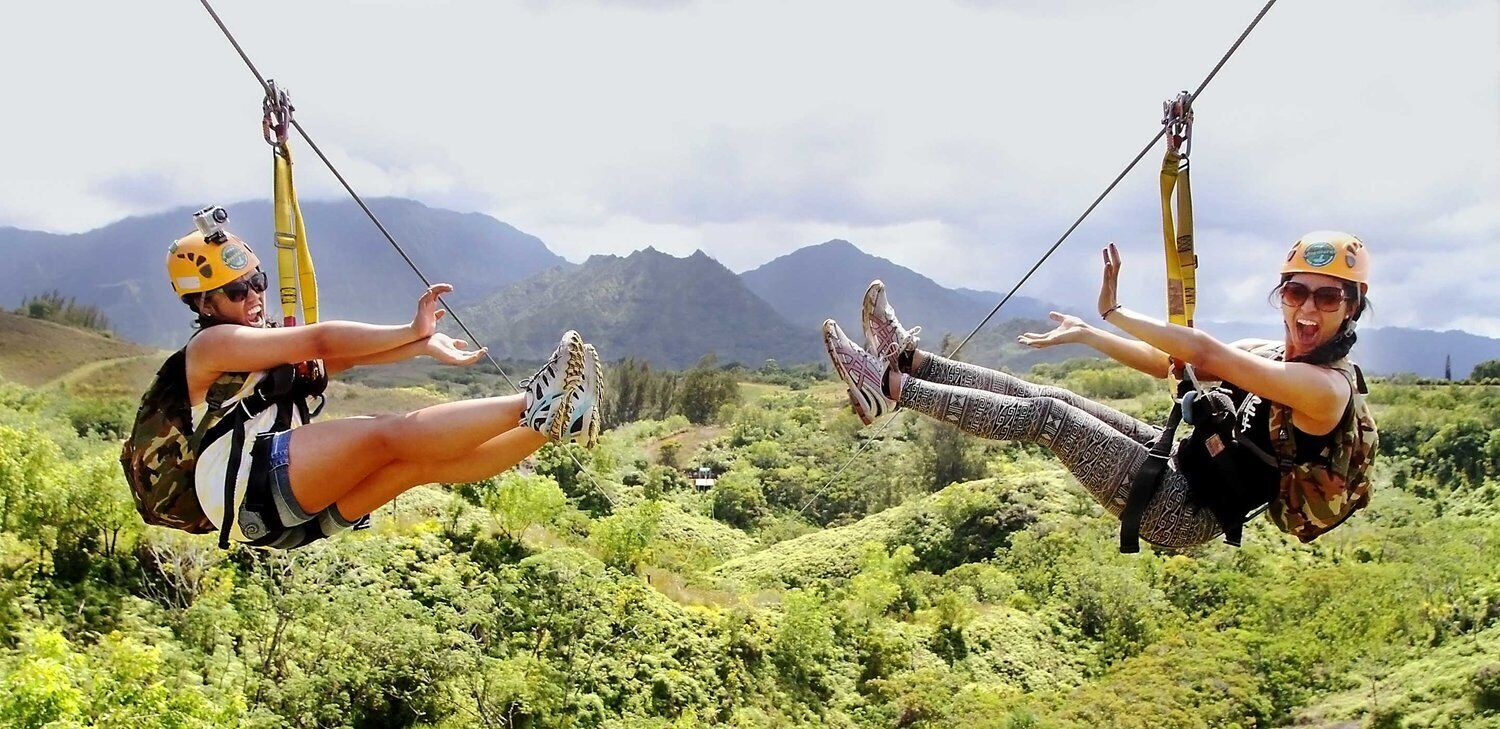 Zwei Personen beim Ziplining vor einer gebirgigen Landschaft.