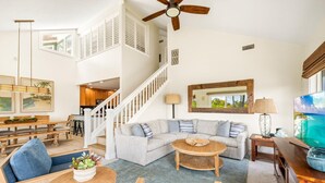 Un vaste salon comprenant un grand canapé d’angle, une table basse ronde et un coin repas avec des meubles en bois. Un escalier mène à l’étage supérieur, et on peut apercevoir un ventilateur de plafond.