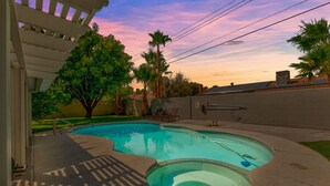 A swimming pool with a hot tub, surrounded by a patio area with a pergola, palm trees, and a brick wall.