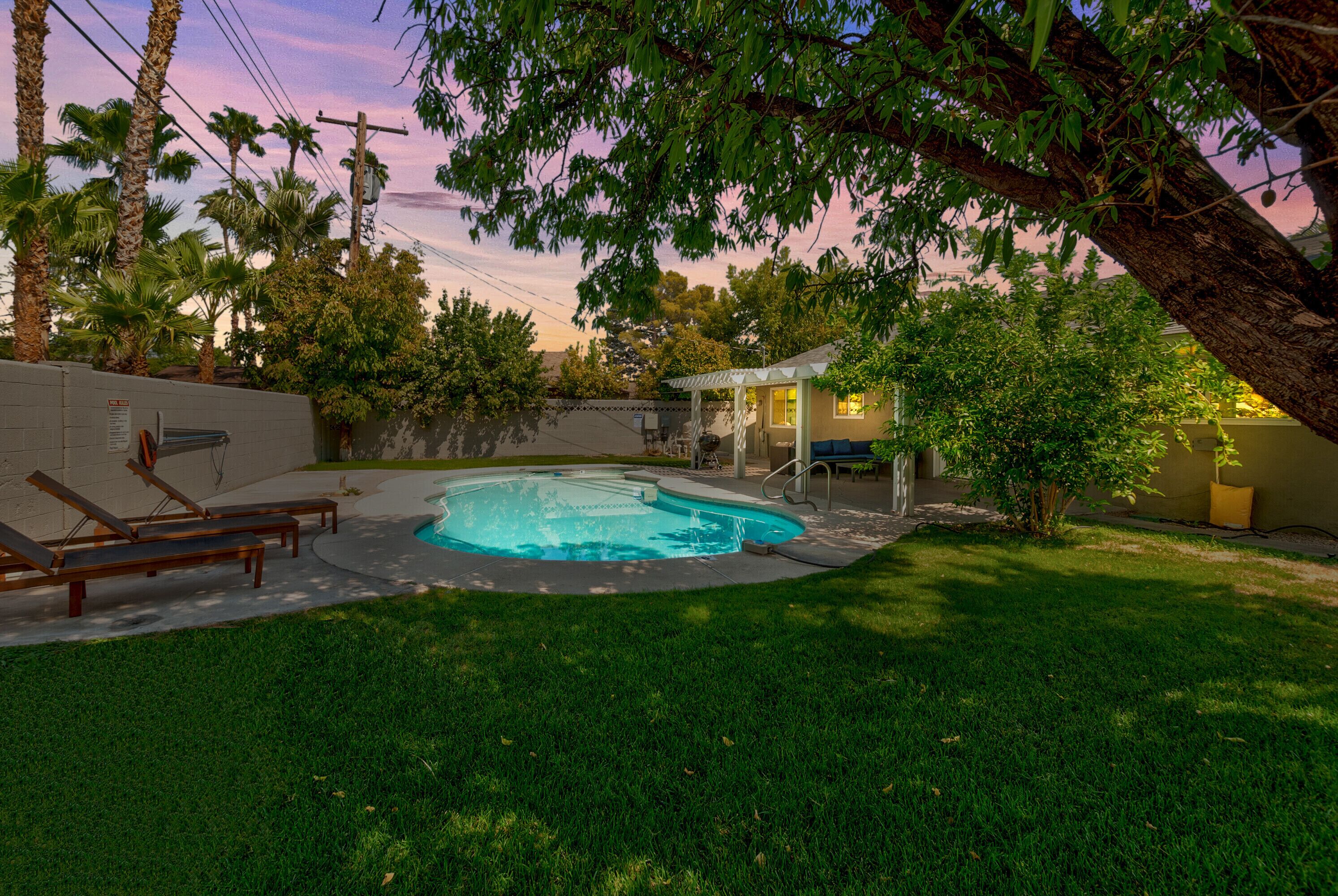 A backyard with a swimming pool, a covered patio, and a lawn with lounge chairs.