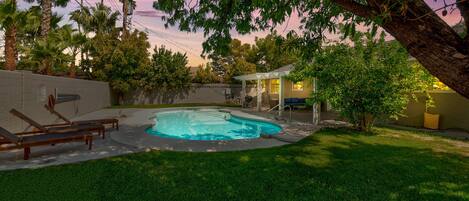 A backyard with a swimming pool, a covered patio, and a lawn with lounge chairs.