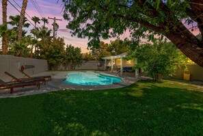 A backyard with a swimming pool, a covered patio, and a lawn with lounge chairs.