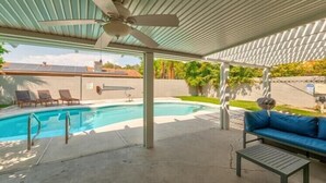 A covered outdoor swimming pool area with a ceiling fan, lounge chairs, and a blue sofa.