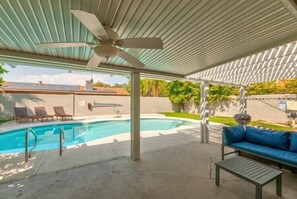 A covered outdoor swimming pool area with a ceiling fan, lounge chairs, and a blue sofa.