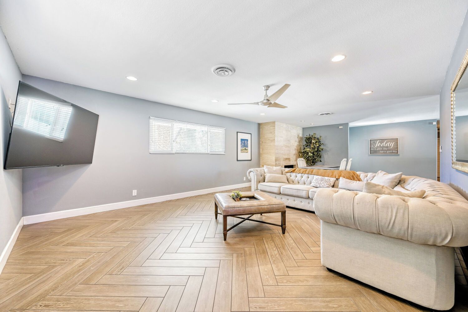 A spacious living room with a herringbone wood floor, a large beige sofa, a coffee table, a flat-screen TV mounted on the wall, and a ceiling fan.