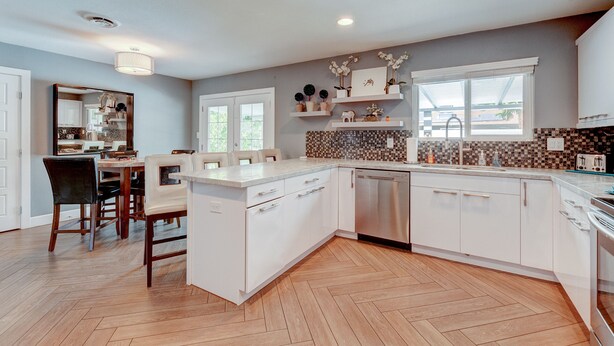 Une cuisine moderne avec des meubles blancs, un parquet en bois disposé en chevrons et un coin repas comprenant une table et des chaises en bois.