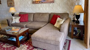 Uma sala de estar com sofá de canto, duas mesas laterais com abajures, uma mesa de centro de vidro e um tapete estampado.