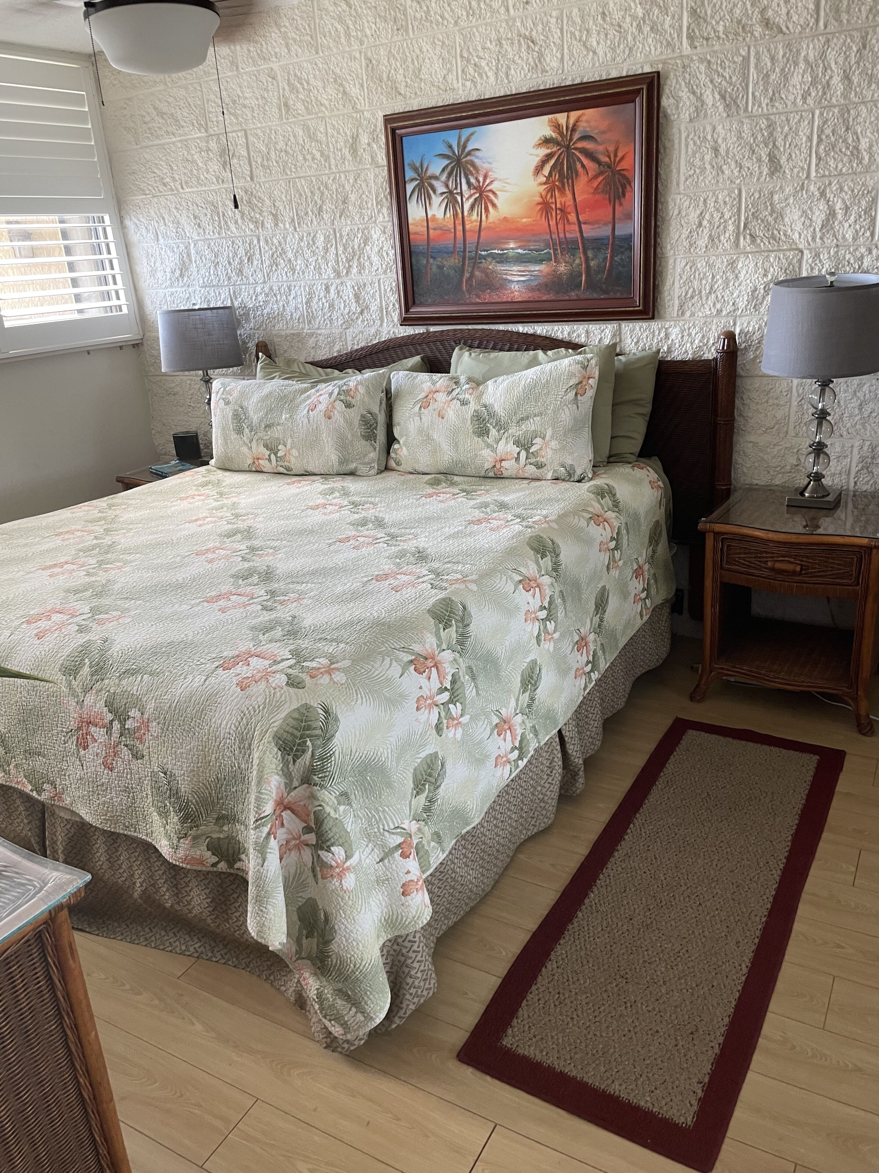 A bedroom with a large bed, a painting of palm trees and a sunset, a nightstand with a lamp, and a wooden floor.