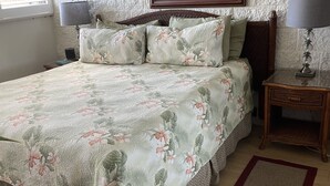 Quarto com uma cama grande, uma pintura de palmeiras e um pôr do sol, mesa de cabeceira com abajur e piso de madeira.