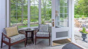 A sunroom with large windows, two patterned chairs, a small table, and a door leading outside. - A Historic Place to Stay for Your Historic Days. Gracious Granite Home + Tavern (Waterford)