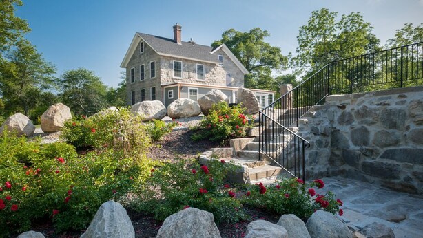 A stone house with a stone staircase and a garden with large rocks and red flowers. - A Historic Place to Stay for Your Historic Days. Gracious Granite Home + Tavern (Waterford)