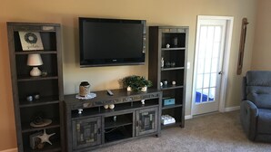 A living room with a TV mounted on the wall, a grey armchair, and a wooden entertainment center with shelves and drawers.