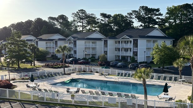 A hotel with a swimming pool, lounge chairs, and a fenced parking area.