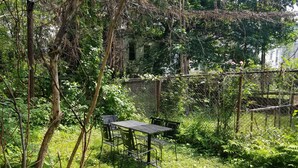 A table and chairs set in a garden with a chain-link fence and dense foliage. - Elegant NYC Pad with Wifi (Queens)
