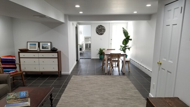 A modern living room with a dark wood coffee table, a white sofa, and a wooden cabinet with framed pictures. A dining area with a wooden table and chairs is visible in the background. - Elegant NYC Pad with Wifi (Queens)