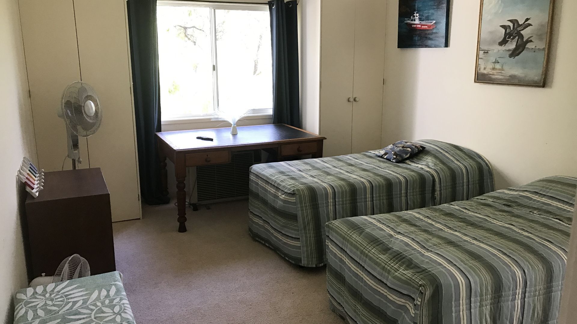 A small, tidy room with two single beds, a desk with a chair, a standing fan, and a window with curtains.