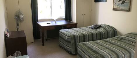 A small, tidy room with two single beds, a desk with a chair, a standing fan, and a window with curtains.