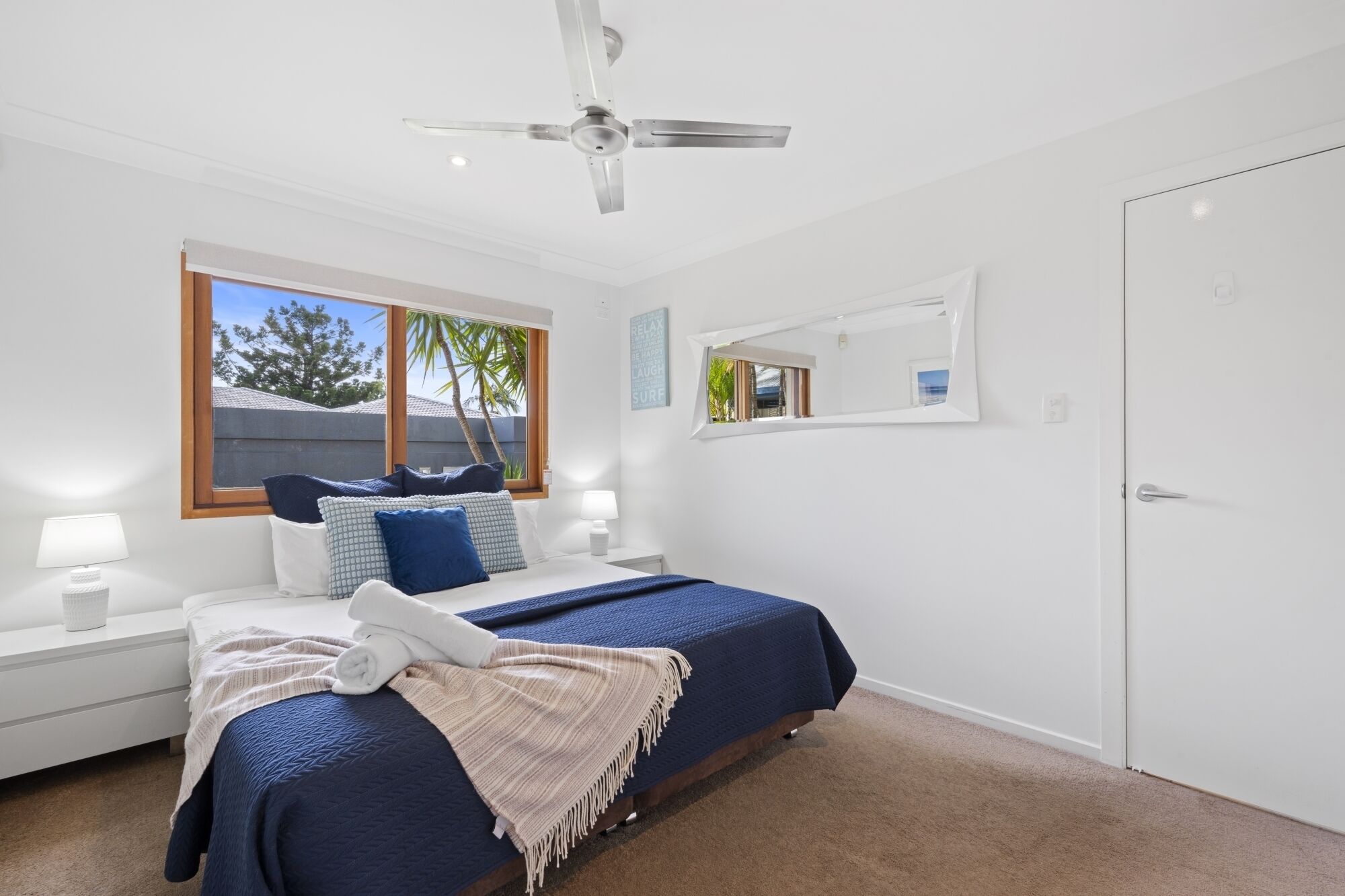 A bedroom with a bed, a ceiling fan, a window with a view, a bedside table with a lamp, and a door.