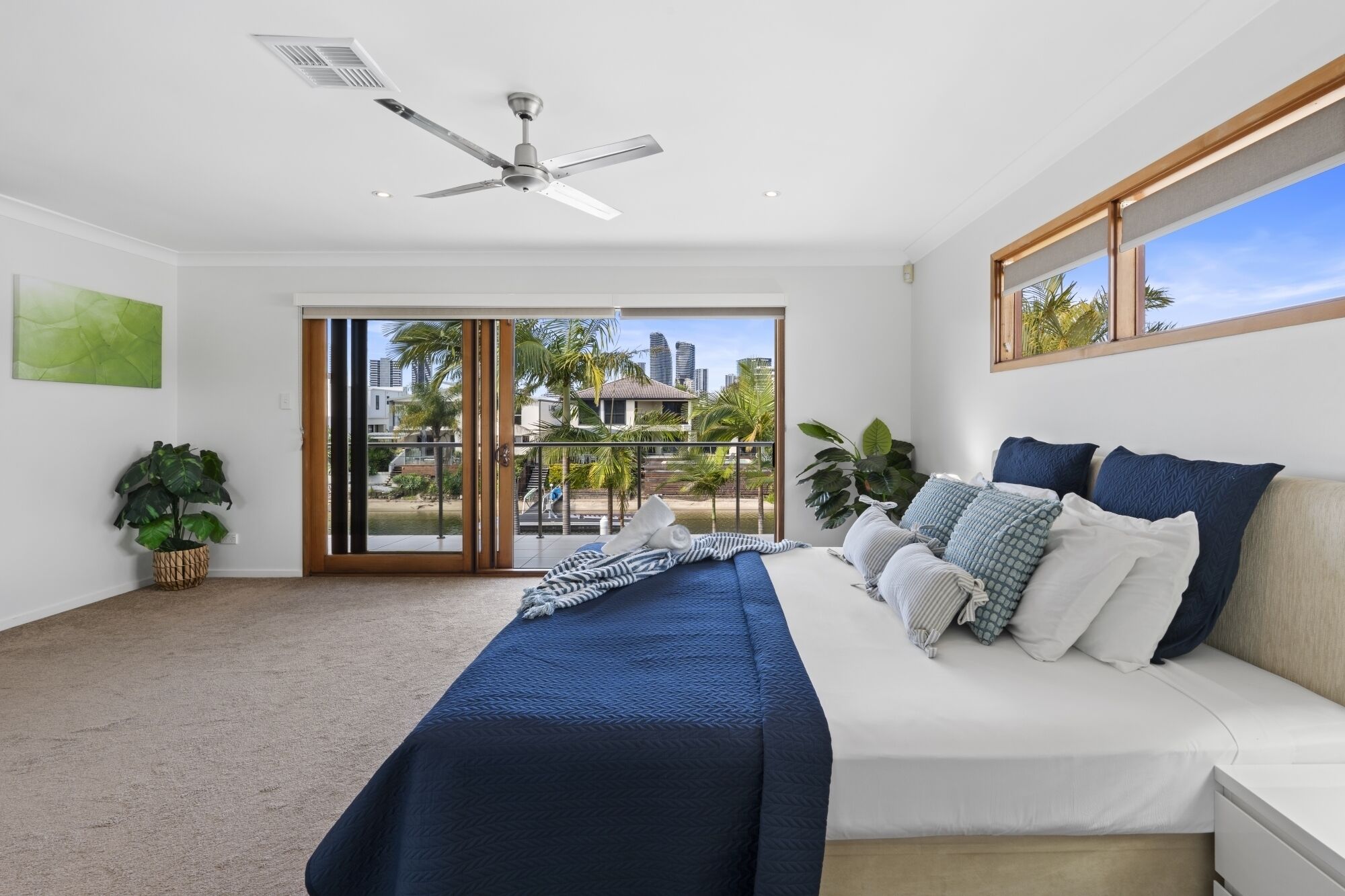 Quarto moderno com cama, ventilador de teto, vista através de uma porta de vidro deslizante para uma varanda, e paisagem urbana ao fundo.