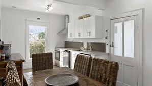 A modern kitchen with white cabinetry, a dining table with wicker chairs, and a large window with a view of greenery. - Healesville Garden Accommodation “GRANDVIEW”   (HEALESVILLE)